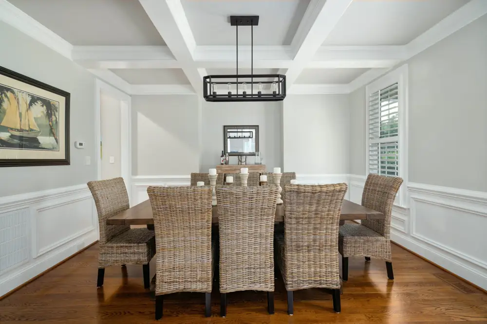 A dining room with a coffered ceiling, white wainscoting, a long wooden table and woven chairs on oak floors.