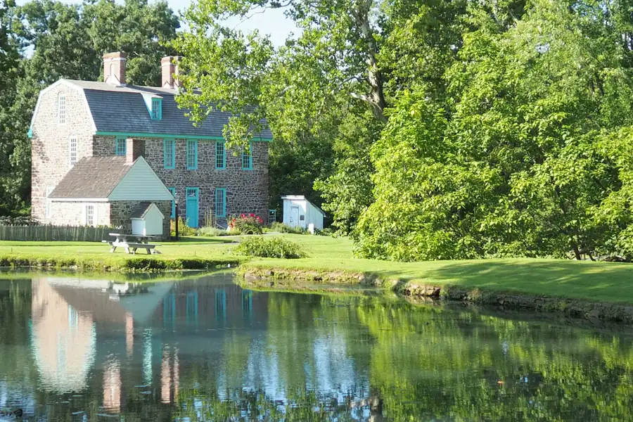An old fieldstone house with a gambrel roof and teal trim beside a still pond that mirrors it, ringed by full summer trees.