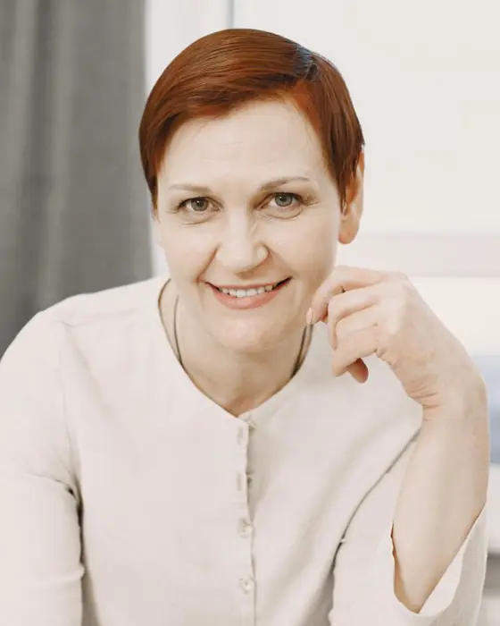 A smiling woman in her fifties with short red hair and a cream linen top, resting her chin on one hand in a bright room.