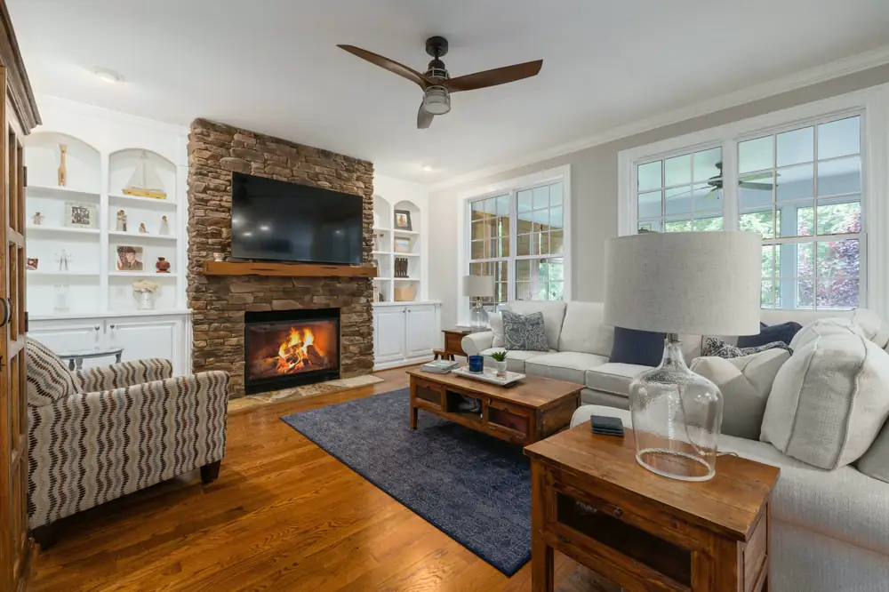 A living room with a stacked stone fireplace, built-in white shelves, a cream sectional and oak floors under a ceiling fan.