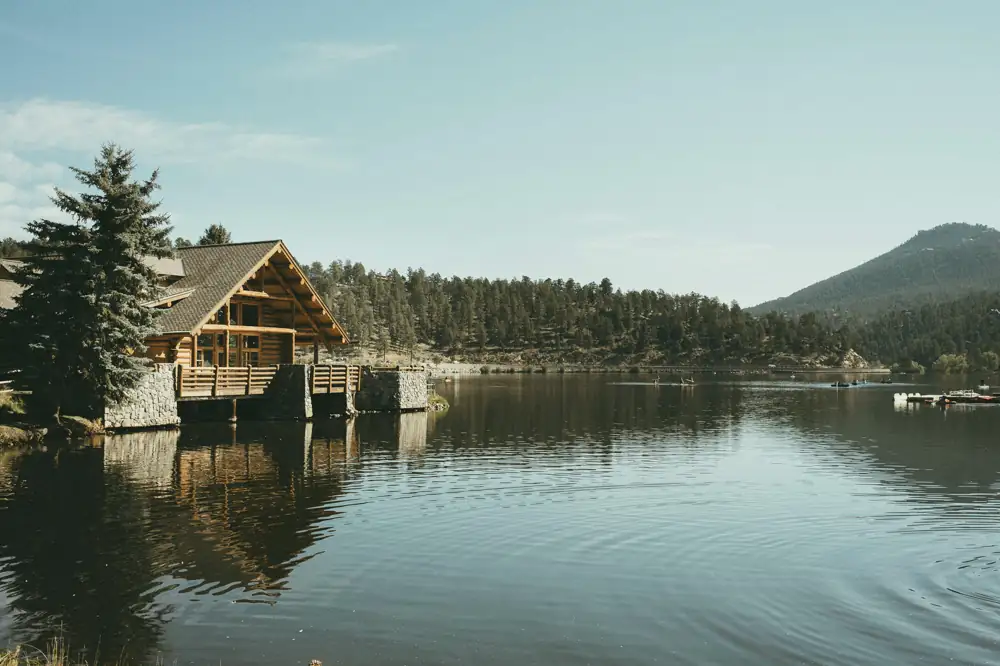 A large log lodge with a timber gable and stone base standing at the edge of a still mountain lake, with pines and a forested hill behind.