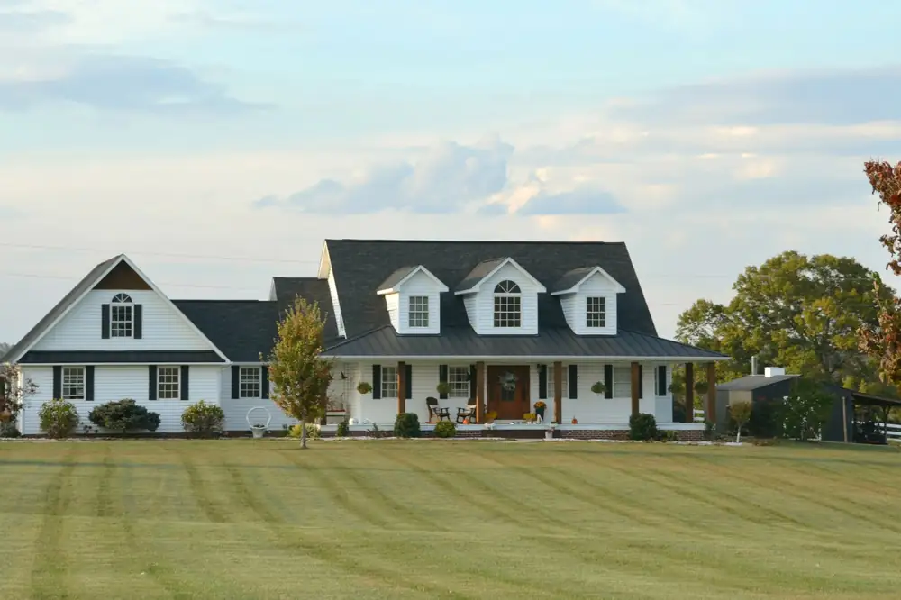 A long white farmhouse with a dark standing seam roof and three dormers across a wide mown lawn, with a dark outbuilding at the right.