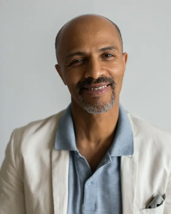 A smiling man with a trimmed silver beard in a cream blazer over a blue polo, against a pale grey wall.
