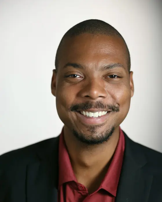 A smiling man with a short beard in a black blazer and a deep red shirt, against a soft white backdrop.