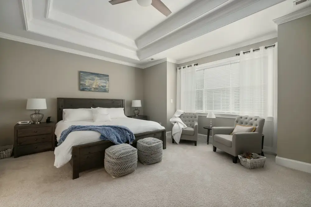 A primary bedroom with a tray ceiling, a dark wood bed with a blue throw, two armchairs and a wide window with sheer curtains.
