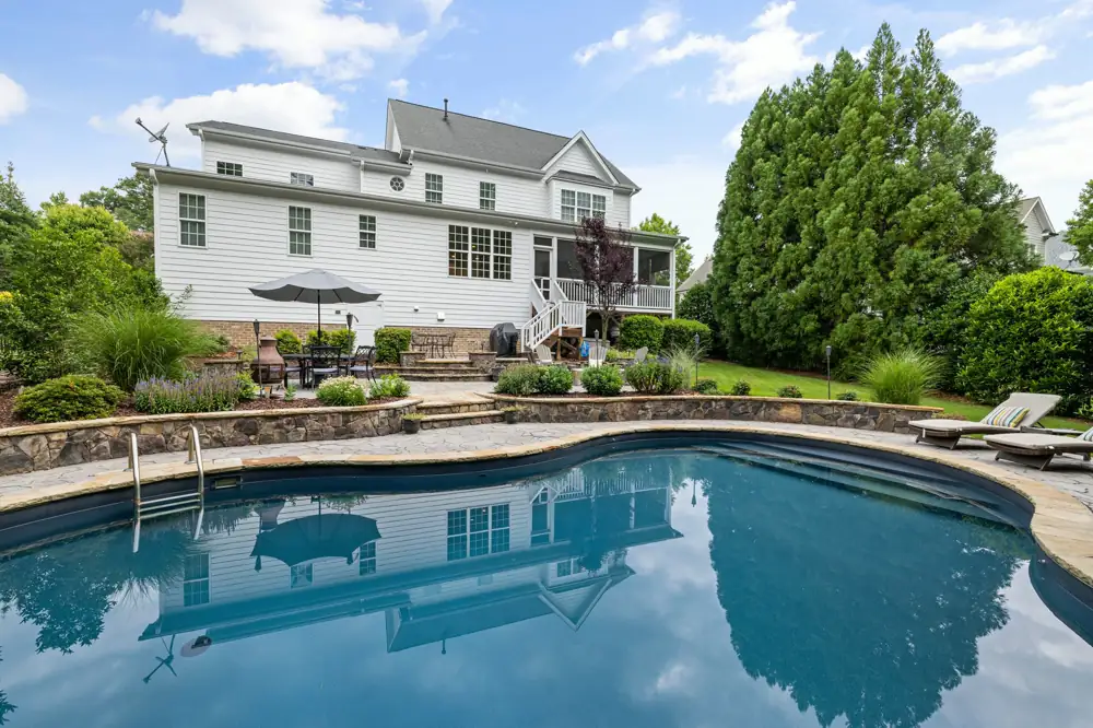 The back of a large white clapboard house with a screened porch, above a curved pool and a stone terrace with a patio table and umbrella.