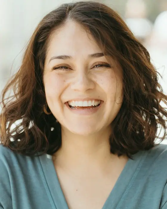 A laughing woman with wavy shoulder-length brown hair in a sage green cardigan, in bright daylight.
